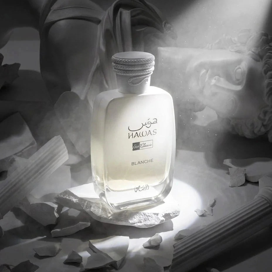 A bottle of HALAS Blanc de Blancs perfume is centered in a dramatic monochrome scene with classical statue fragments and a faint statue face in the background. The bottle is frosted white with dark lettering, and a spotlight illuminates the front of the bottle.