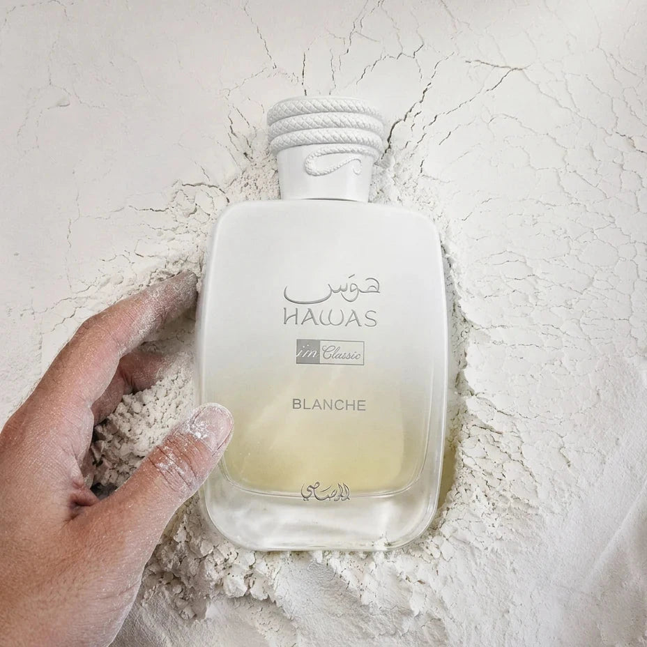 A hand holds a frosted glass perfume bottle labeled 'HUMAIAS NALUA BLANCHE' against a textured, cracked off-white background with white powder scattered around it.