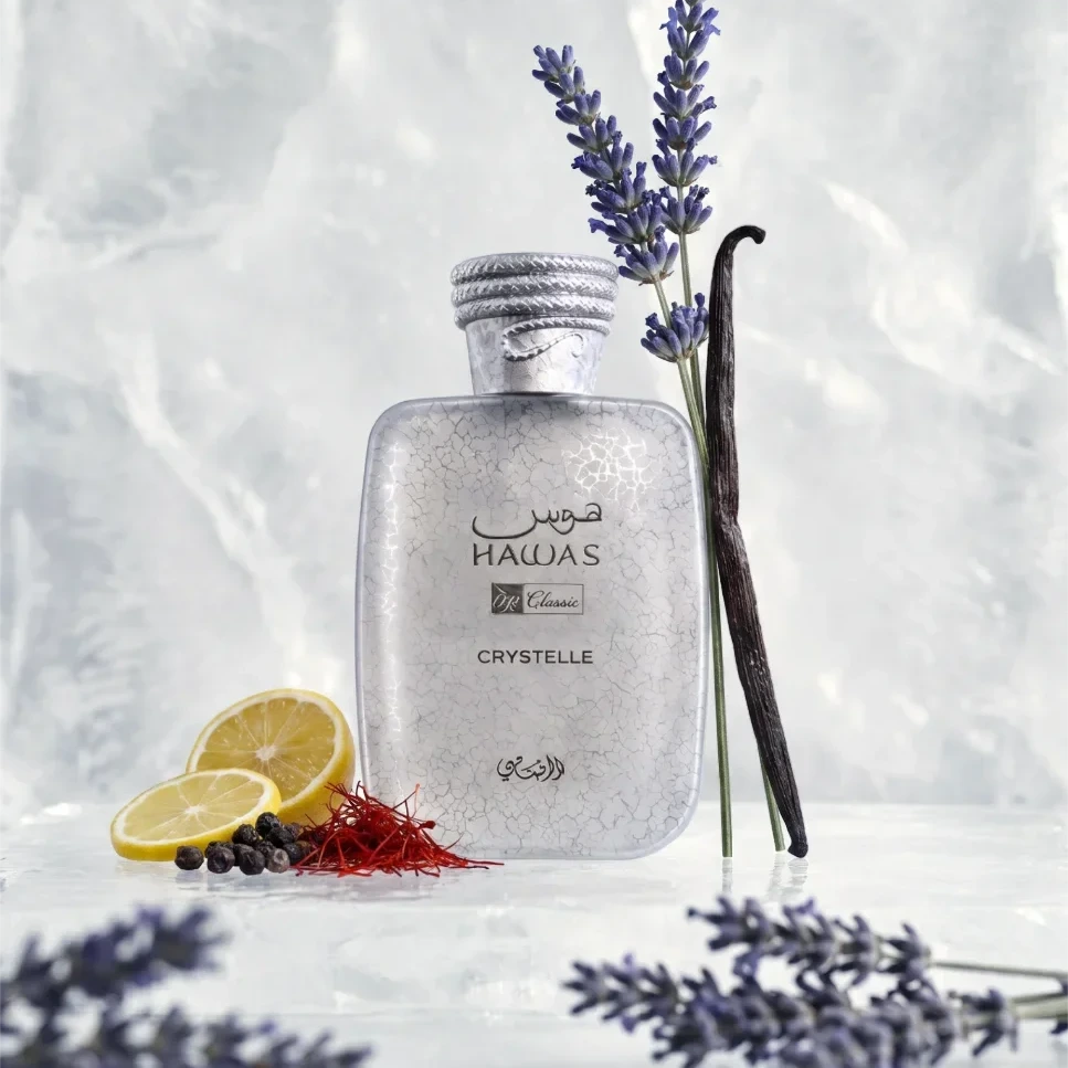 A frosted glass bottle labeled HAULAS Classic Crystelle stands on a light surface with lavender sprigs, a vanilla bean, lemon slices, and saffron threads around it against a soft marble-like background.